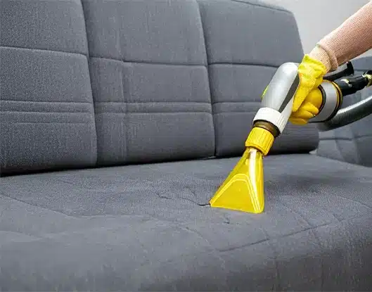 A cleaning technician's hands holding a vacuum cleaner while using it on a sofa upholstery.