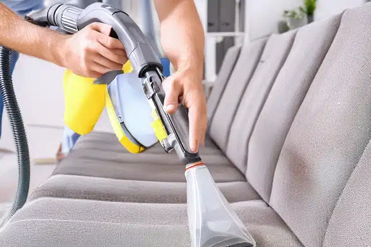 A cleaning technician's hand is using a damp cloth on a sofa upholstery in a living room.