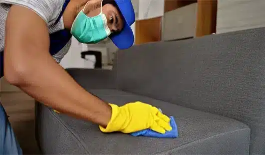 A cleaning technician, wearing a uniform and a face mask, is using a special sponge to clean the sofa upholstery in a living room.